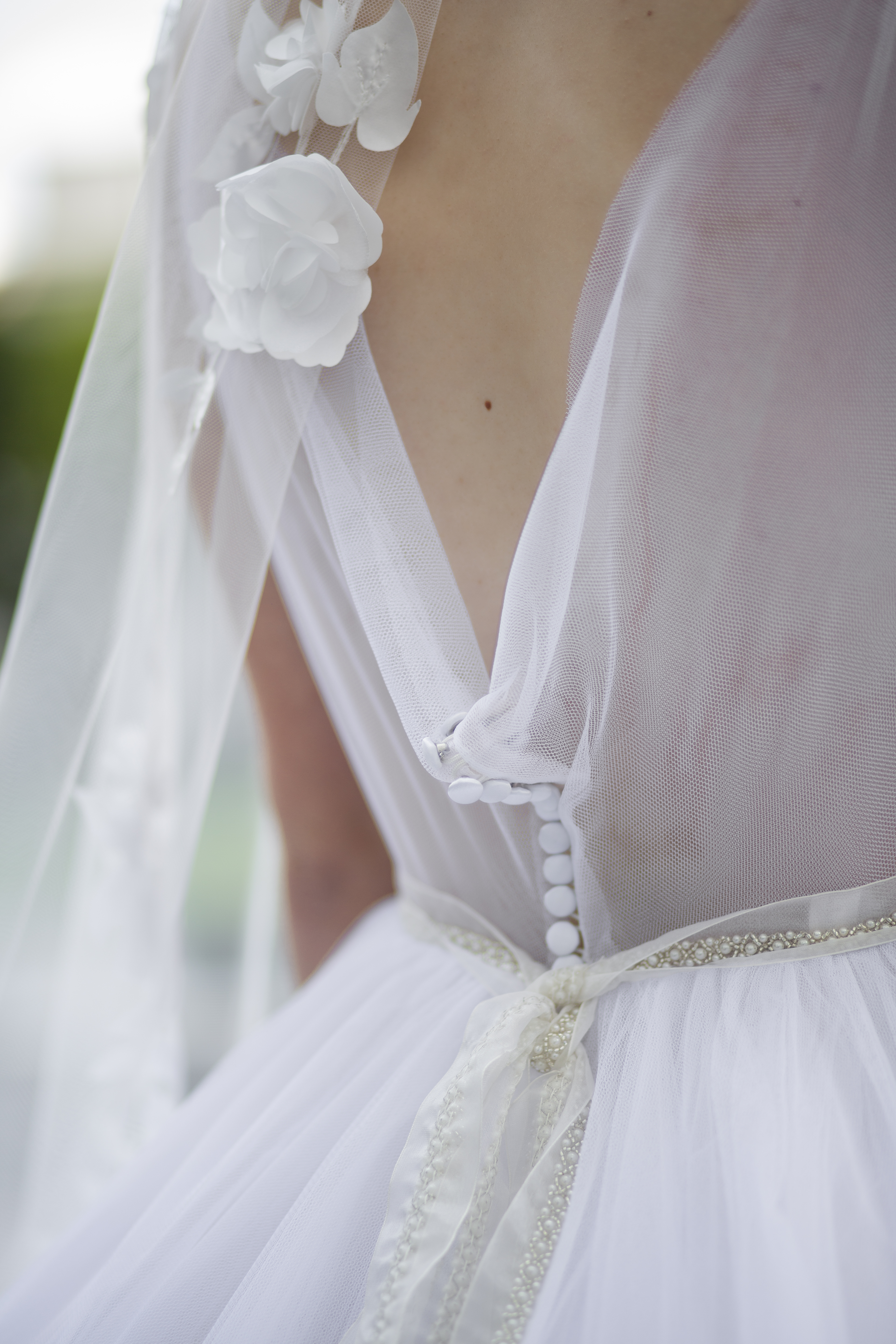 A close up photo of the back of a bride wearing a bridal dress and romantic floral calf length veil. The frame of the image begins at her upper shoulder blades and finishes at her mid back. You can see the tulle cowl neckline of the gown with the veil flowing across the left side of the image. On the veil are large roses with leafs above and below the flowers. You can see one flower prominently on the edge of the veil with a leaf and stem above. You can see some stitching on the leaf that is designed to look like veining on the leaf. The petals and leaf are 3 dimensional.